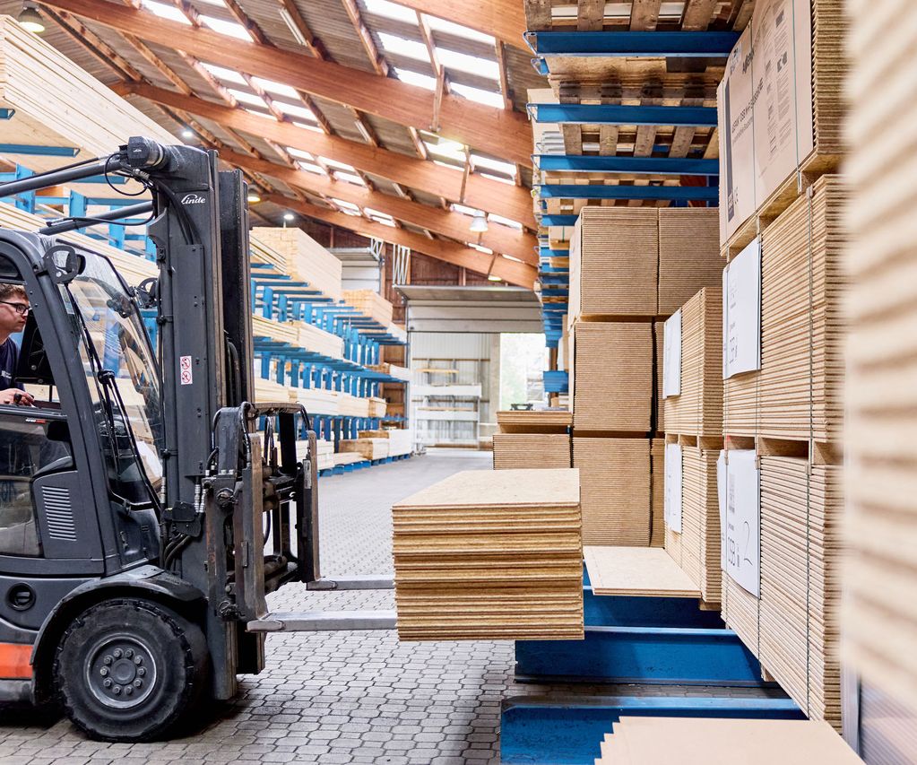 Ein Gabelstaplerfahrer transportiert gestapelte OSB Platten in einem Lagerhaus mit hohen Regalen und einer Holzdecke.
