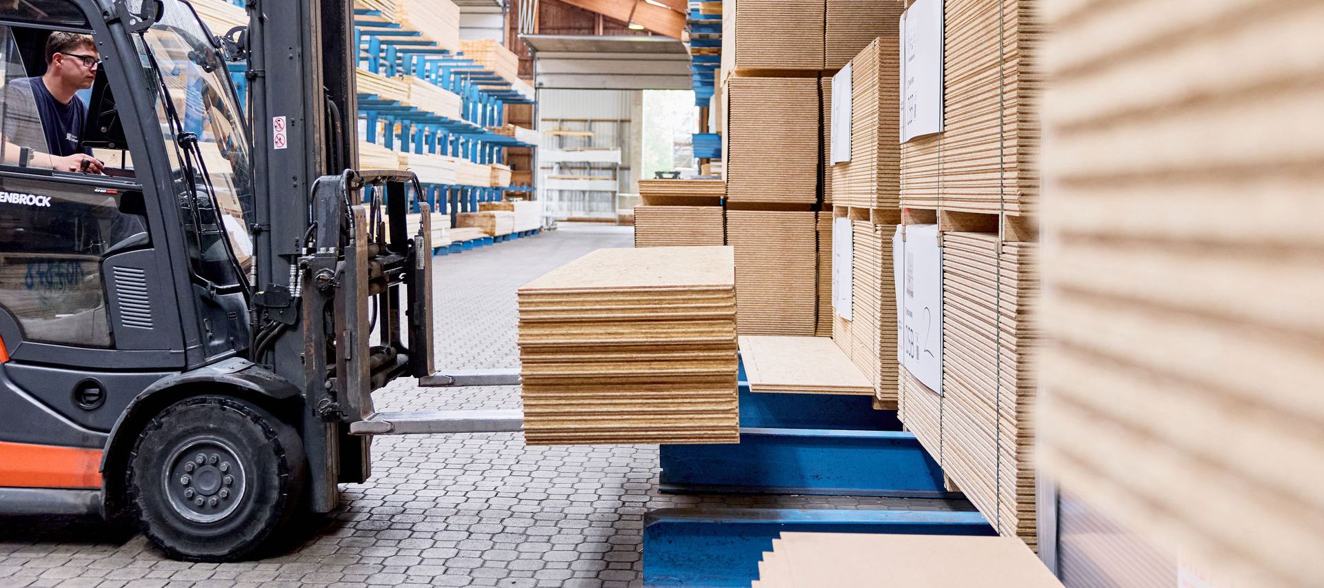 Ein Gabelstaplerfahrer transportiert gestapelte OSB Platten in einem Lagerhaus mit hohen Regalen und einer Holzdecke.