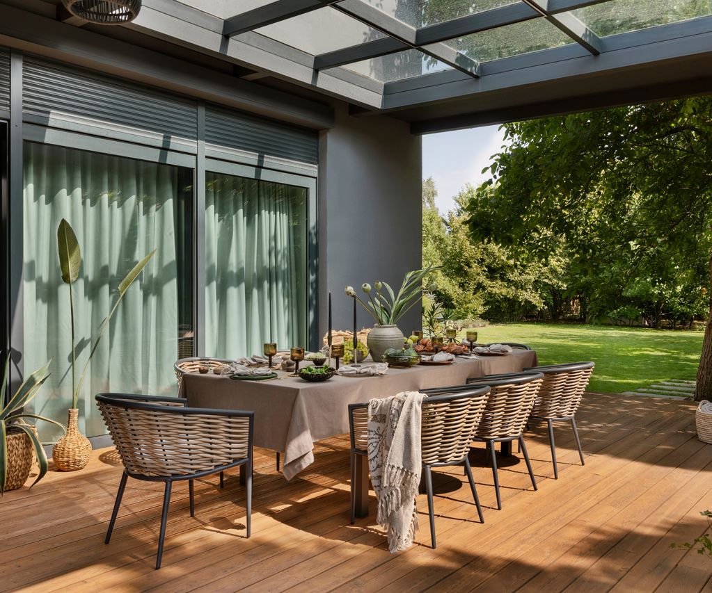 errasse mit braunen Terrassendielen, Esstisch, Rattanstühlen und Glasüberdachung vor einem Garten
