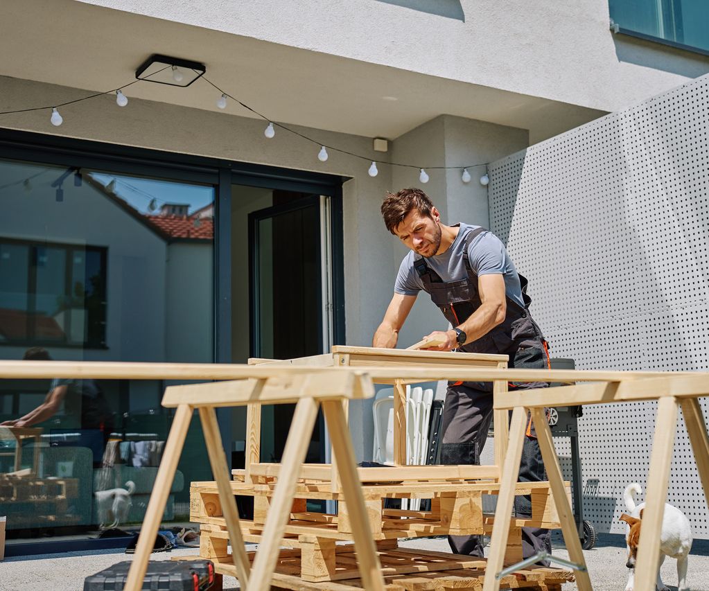 Handwerker bearbeitet Holz auf einem Arbeitsbock auf einer Terrasse vor einem Wohnhaus