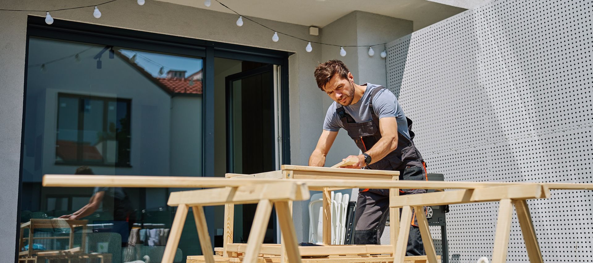 Handwerker bearbeitet Holz auf einem Arbeitsbock auf einer Terrasse vor einem Wohnhaus