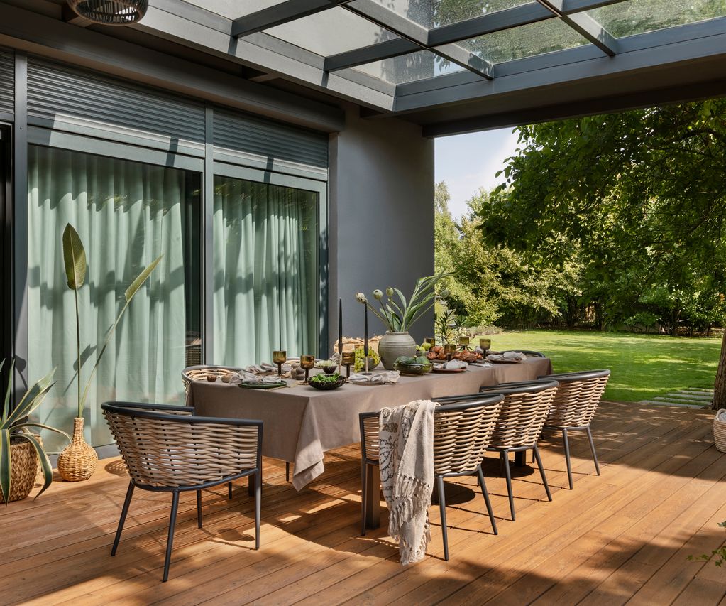 Überdachter Essbereich auf einer Holzterrasse mit gedecktem Tisch, Geflechtstühlen und Blick in den Garten