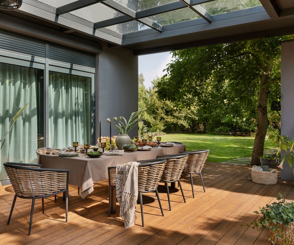 Überdachter Essbereich auf einer Holzterrasse mit gedecktem Tisch, Geflechtstühlen und Blick in den Garten