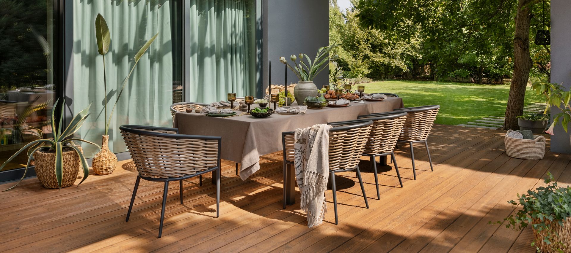 Überdachter Essbereich auf einer Holzterrasse mit gedecktem Tisch, Geflechtstühlen und Blick in den Garten