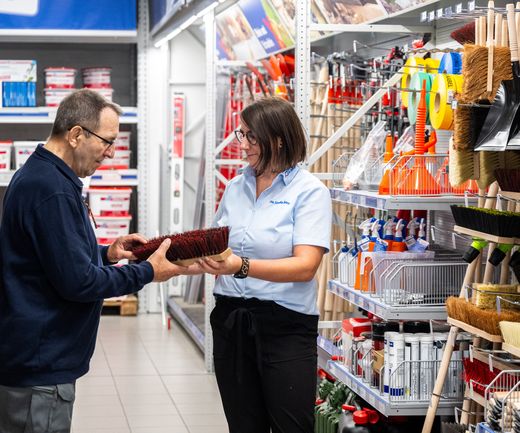 Fachberaterin zeigt Kunde Besen. Sie befinden sich im Handwerkerfachmarkt