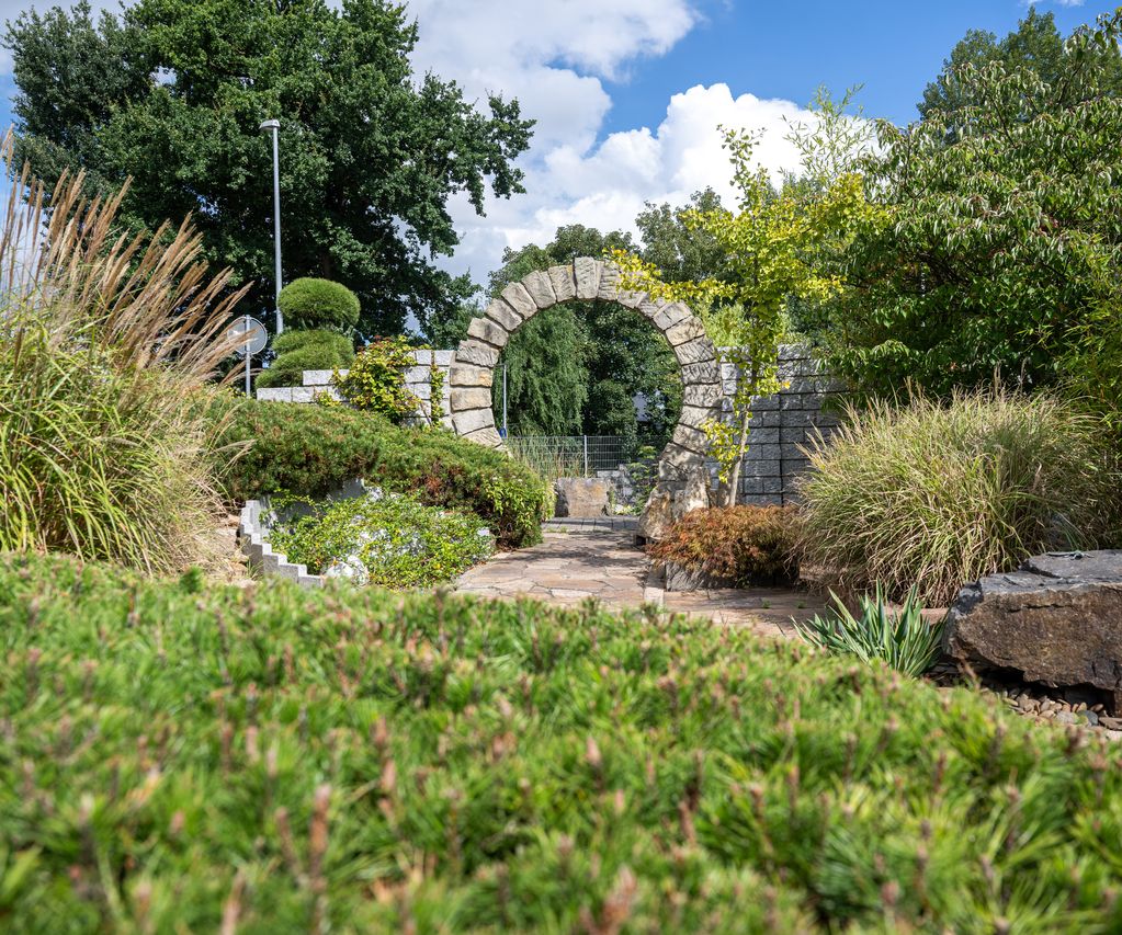 Natursteinbogen in einer Gartenausstellung mit bepflanzten Beeten und geschwungenem Gartenweg