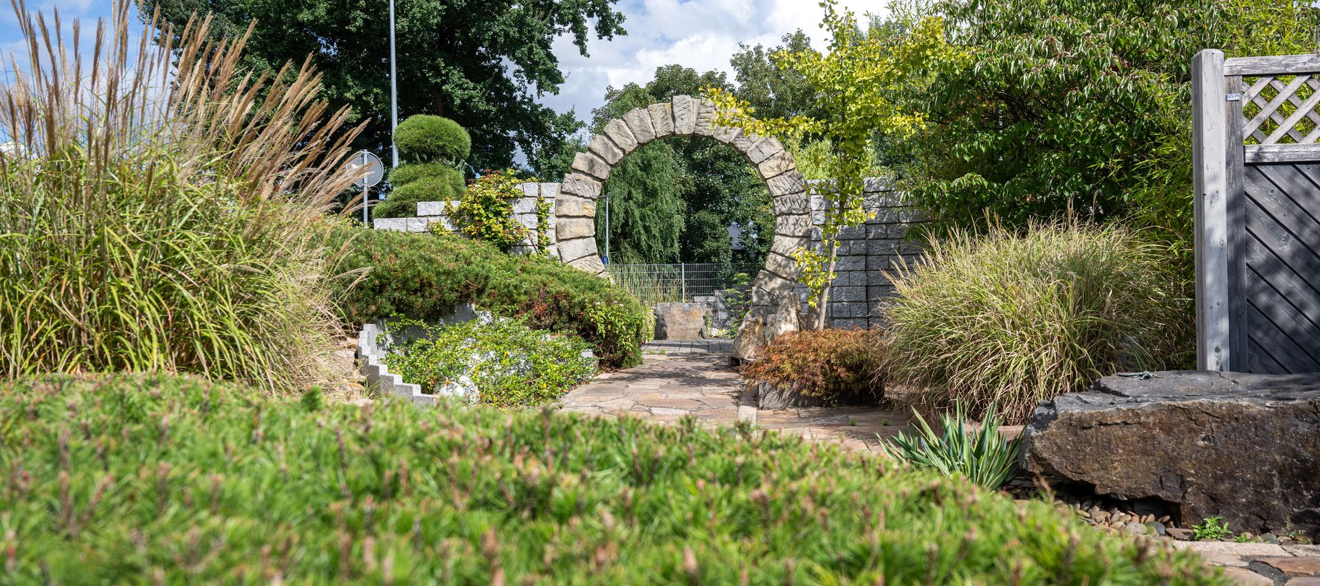 Natursteinbogen in einer Gartenausstellung mit bepflanzten Beeten und geschwungenem Gartenweg