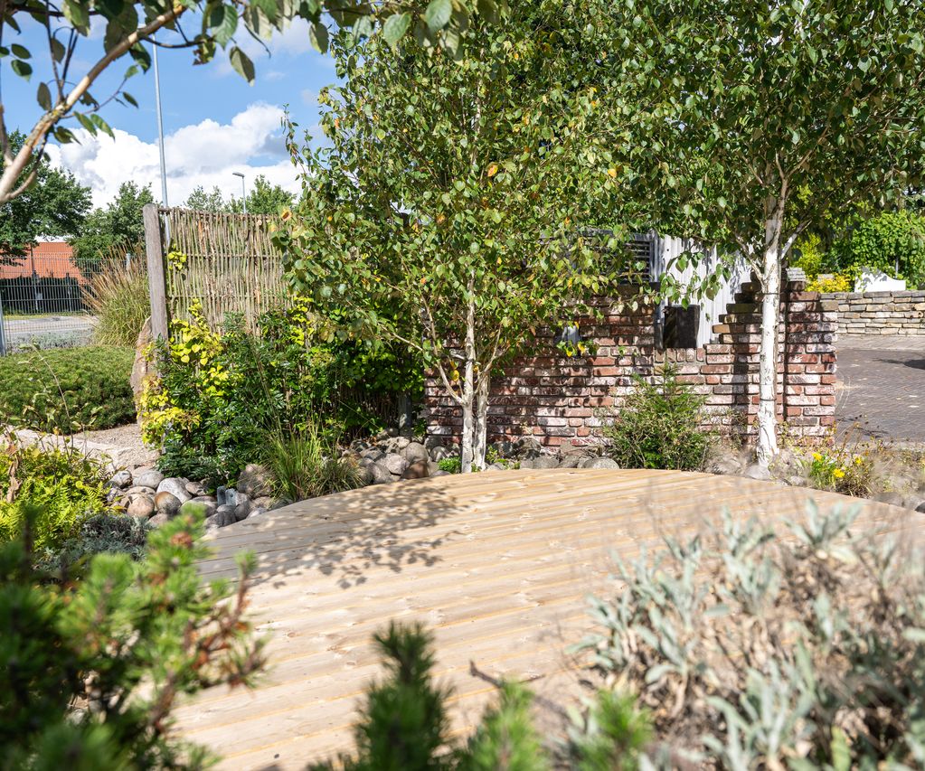 Holzterrasse in einer grünen Gartenausstellung mit Bäumen, Natursteinen und gemauerter Wand
