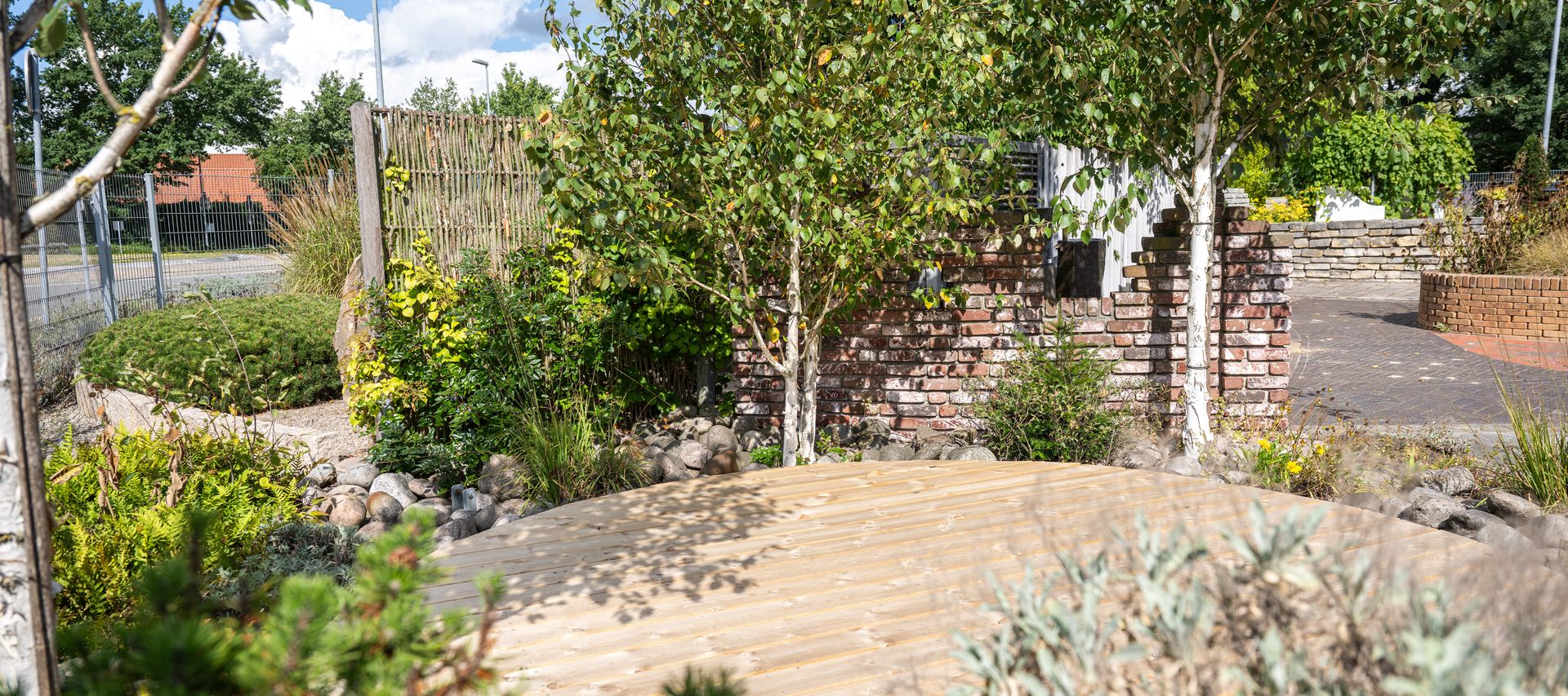 Holzterrasse in einer grünen Gartenausstellung mit Bäumen, Natursteinen und gemauerter Wand