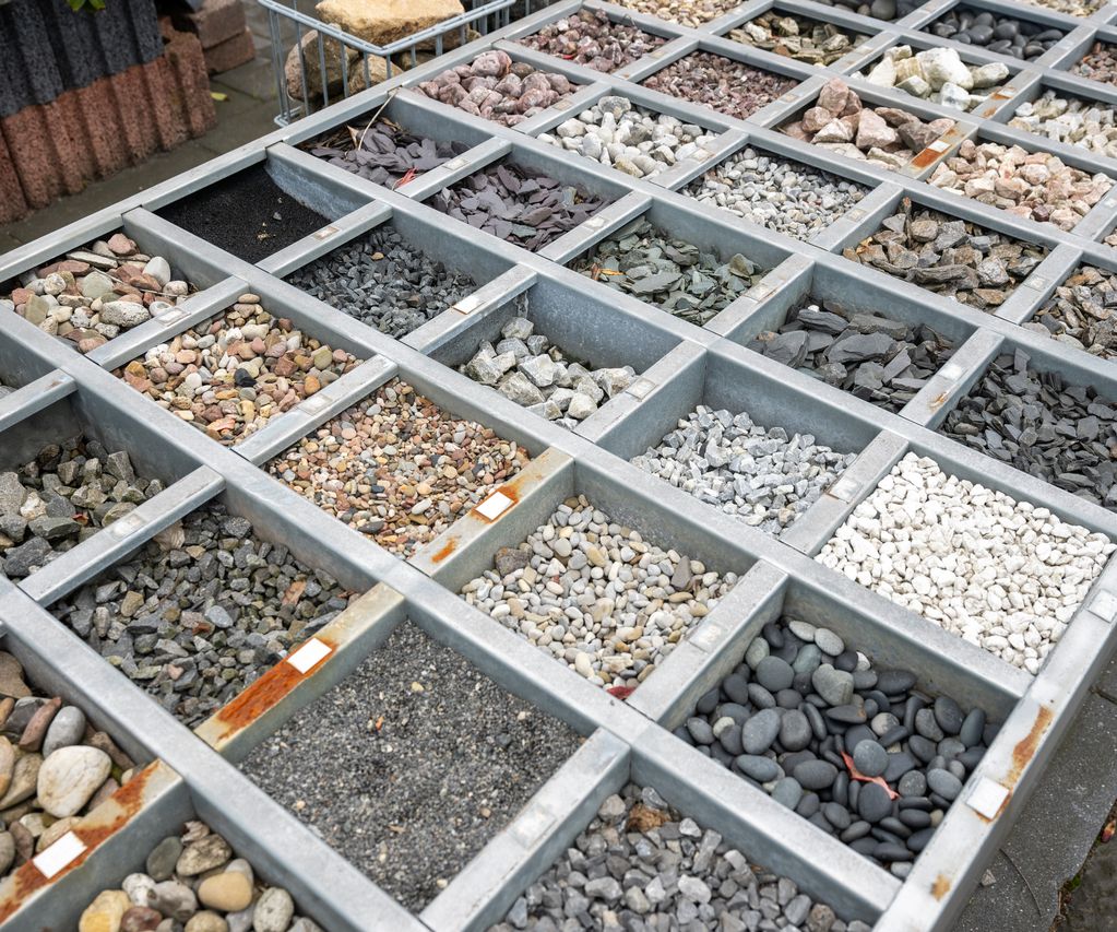 Übersicht einer Musterfläche mit verschiedenen Zierkiesen und Splittsorten in Metallrahmen, ausgelegt zur Material- und Farbauswahl im Außenbereich.