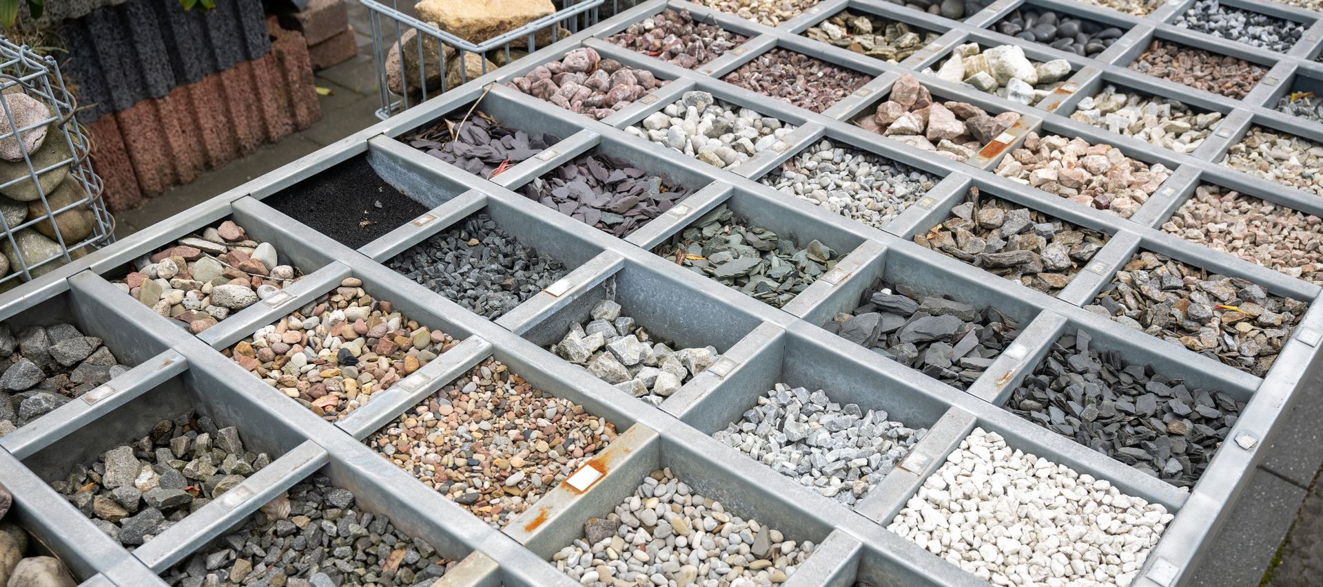 Übersicht einer Musterfläche mit verschiedenen Zierkiesen und Splittsorten in Metallrahmen, ausgelegt zur Material- und Farbauswahl im Außenbereich.