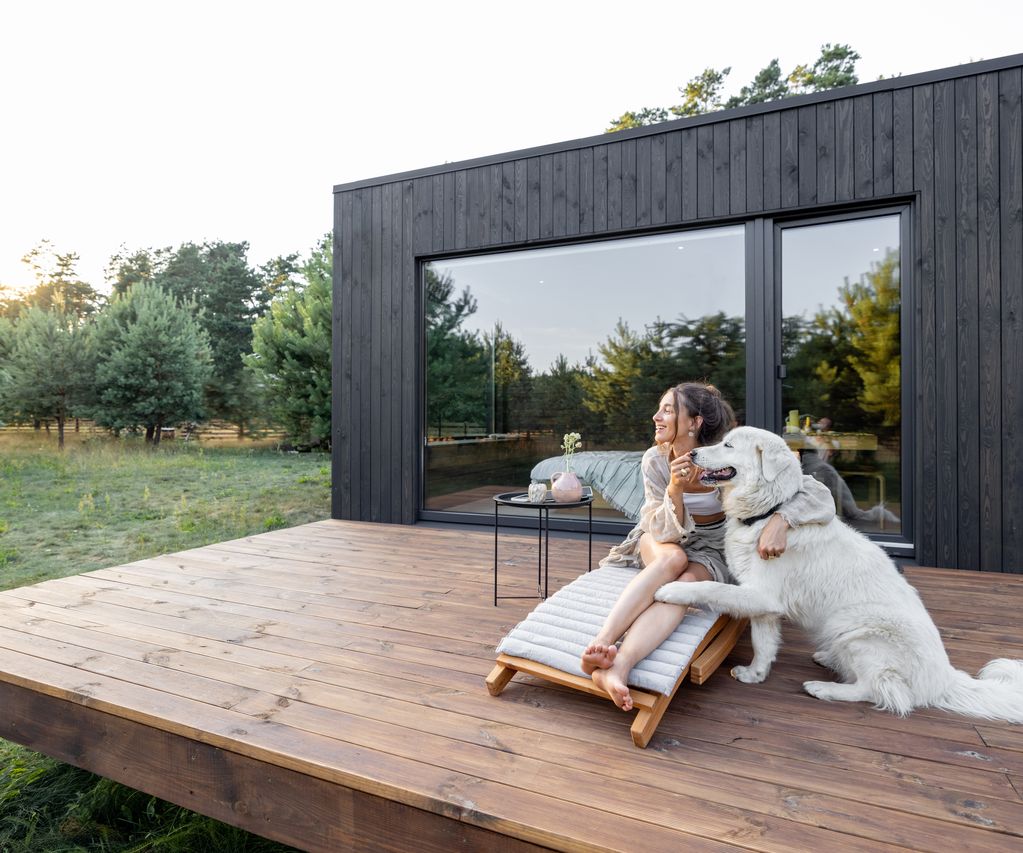 Frau mit weißem Hund auf einer Holzterrasse vor einem dunkel verkleideten Gartenhaus