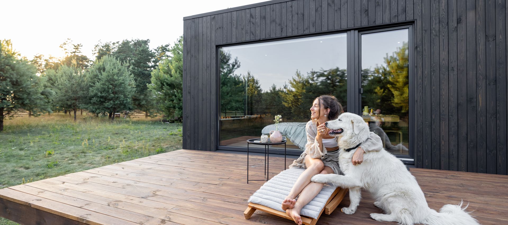Frau mit weißem Hund auf einer Holzterrasse vor einem dunkel verkleideten Gartenhaus