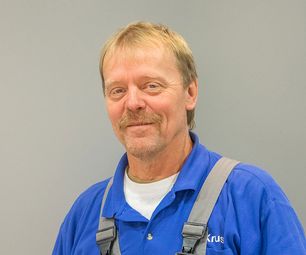 Mitarbeiter von bauXpert steht vor neutralem Hintergrund und blickt freundlich in die Kamera. Er trägt blaue Arbeitskleidung mit Trägern.