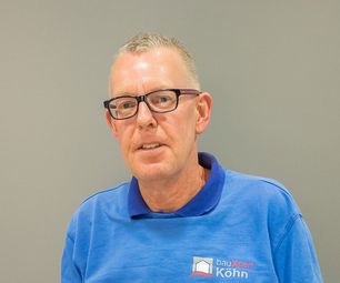 Mitarbeiter von bauXpert steht vor neutralem Hintergrund und blickt aufmerksam in die Kamera. Er trägt blaue Arbeitskleidung mit Firmenlogo und eine Brille.