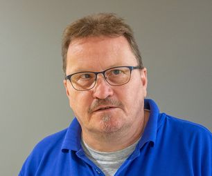 Mitarbeiter von bauXpert mit Brille blickt ruhig in die Kamera. Er trägt ein blaues Poloshirt vor neutralem Hintergrund.