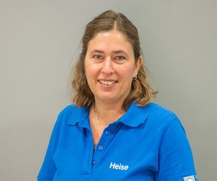 Mitarbeiterin von bauXpert steht vor neutralem Hintergrund und blickt freundlich in die Kamera. Sie trägt blaue Arbeitskleidung mit Namenszug.