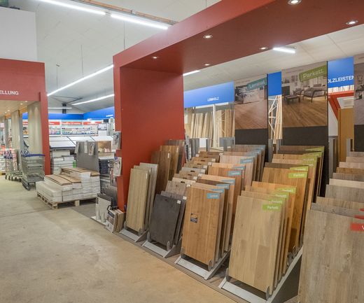Innenaufnahme der Bodenausstellung bei bauXpert mit Musterständern für Laminat, Parkett und Vinyl in verschiedenen Holzoptiken. Im Hintergrund sind weitere Ausstellungs- und Verkaufsbereiche zu sehen.