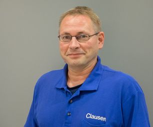 Mitarbeiter von bauXpert steht vor neutralem Hintergrund und blickt freundlich in die Kamera. Er trägt Arbeitskleidung mit Namenszug.