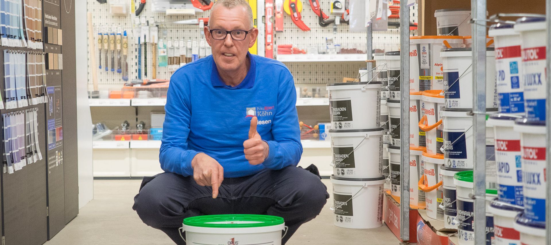 Mitarbeiter von bauXpert kniet im Fachmarkt zwischen Farbreihen und zeigt auf einen Eimer weiße Innenfarbe. Er gibt ein positives Handzeichen und präsentiert das Produkt im Regalgang.