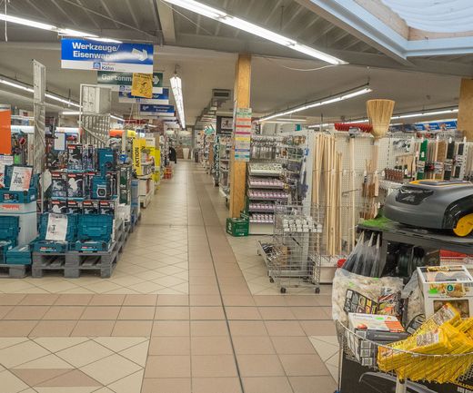 Blick durch den Fachmarkt bei bauXpert mit Regalen für Werkzeuge, Eisenwaren und Gartenbedarf. In den Gängen sind Aktionsflächen und verschiedene Produktpräsentationen aufgebaut.