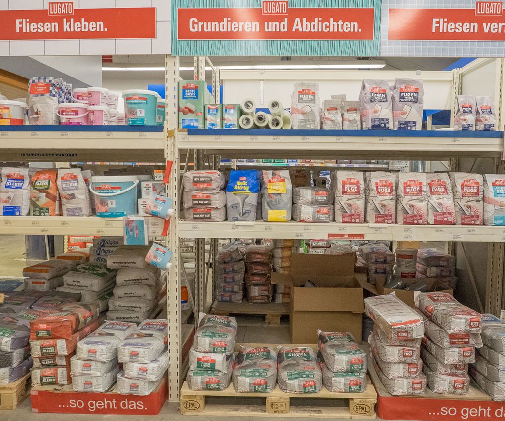 Regalfläche im Fachmarkt mit Bauchemie für Fliesenarbeiten, darunter Kleber, Grundierungen, Abdichtungen und Fugenmörtel in Säcken und Eimern. Die Produkte sind übersichtlich nach Arbeitsschritten sortiert.