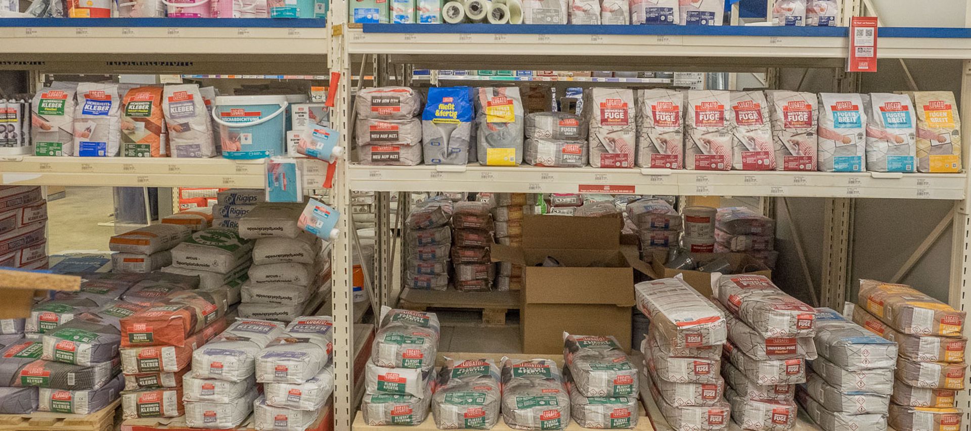 Regalfläche im Fachmarkt mit Bauchemie für Fliesenarbeiten, darunter Kleber, Grundierungen, Abdichtungen und Fugenmörtel in Säcken und Eimern. Die Produkte sind übersichtlich nach Arbeitsschritten sortiert.