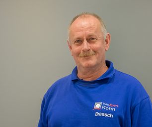 Mitarbeiter von bauXpert steht vor neutralem Hintergrund und blickt freundlich in die Kamera. Er trägt Arbeitskleidung mit Firmenlogo und Namenszug.