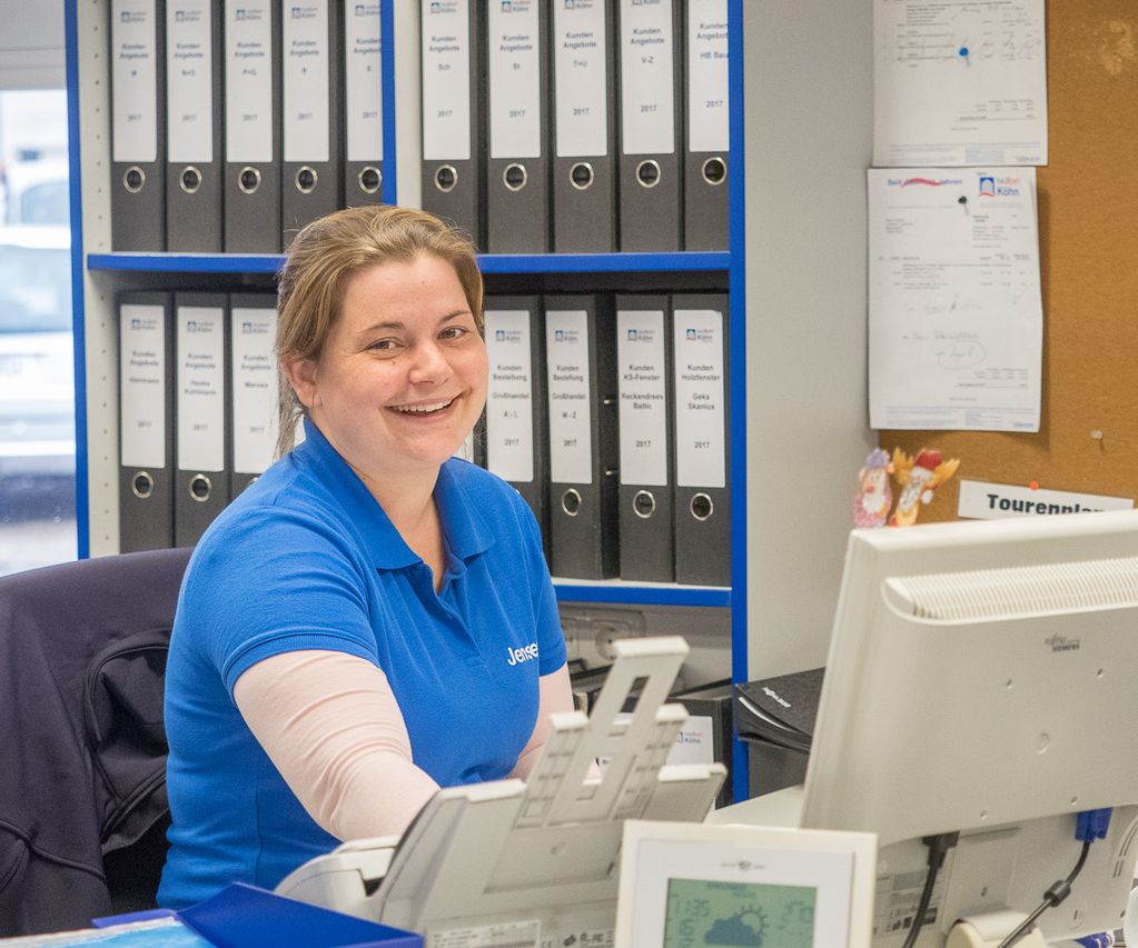 Mitarbeiterin sitzt am Schreibtisch im Büro bei bauXpert und lächelt in die Kamera, im Hintergrund stehen Aktenordner und eine Pinnwand. Auf dem Tisch befinden sich Computer, Telefon und Unterlagen.
