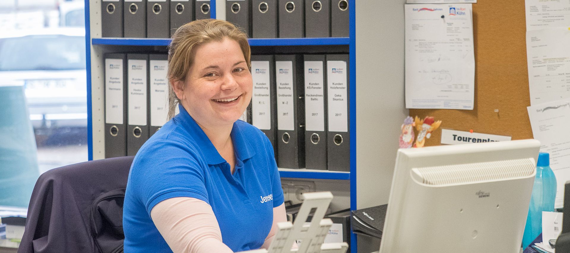 Mitarbeiterin sitzt am Schreibtisch im Büro bei bauXpert und lächelt in die Kamera, im Hintergrund stehen Aktenordner und eine Pinnwand. Auf dem Tisch befinden sich Computer, Telefon und Unterlagen.