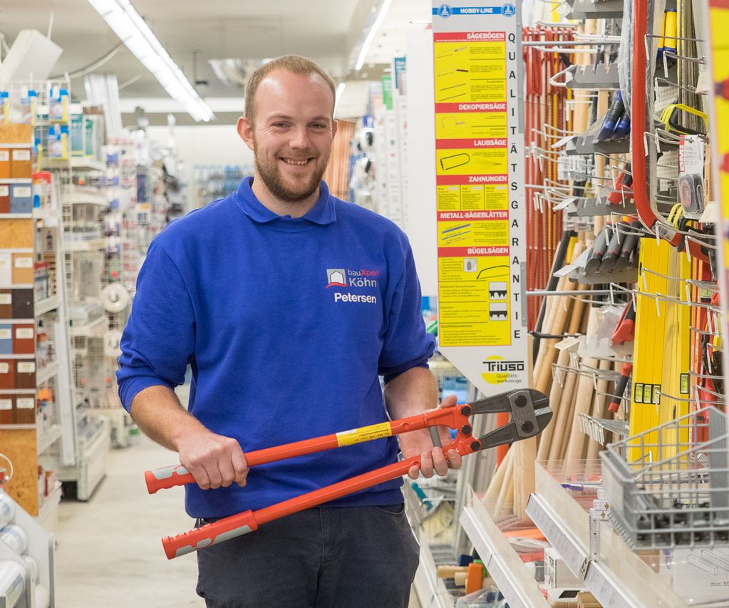 Mitarbeiter von bauXpert steht im Fachmarkt im Werkzeuggang und hält eine Bolzenschere in den Händen. Im Hintergrund sind Regale mit Handwerkzeugen und Zubehör zu sehen.