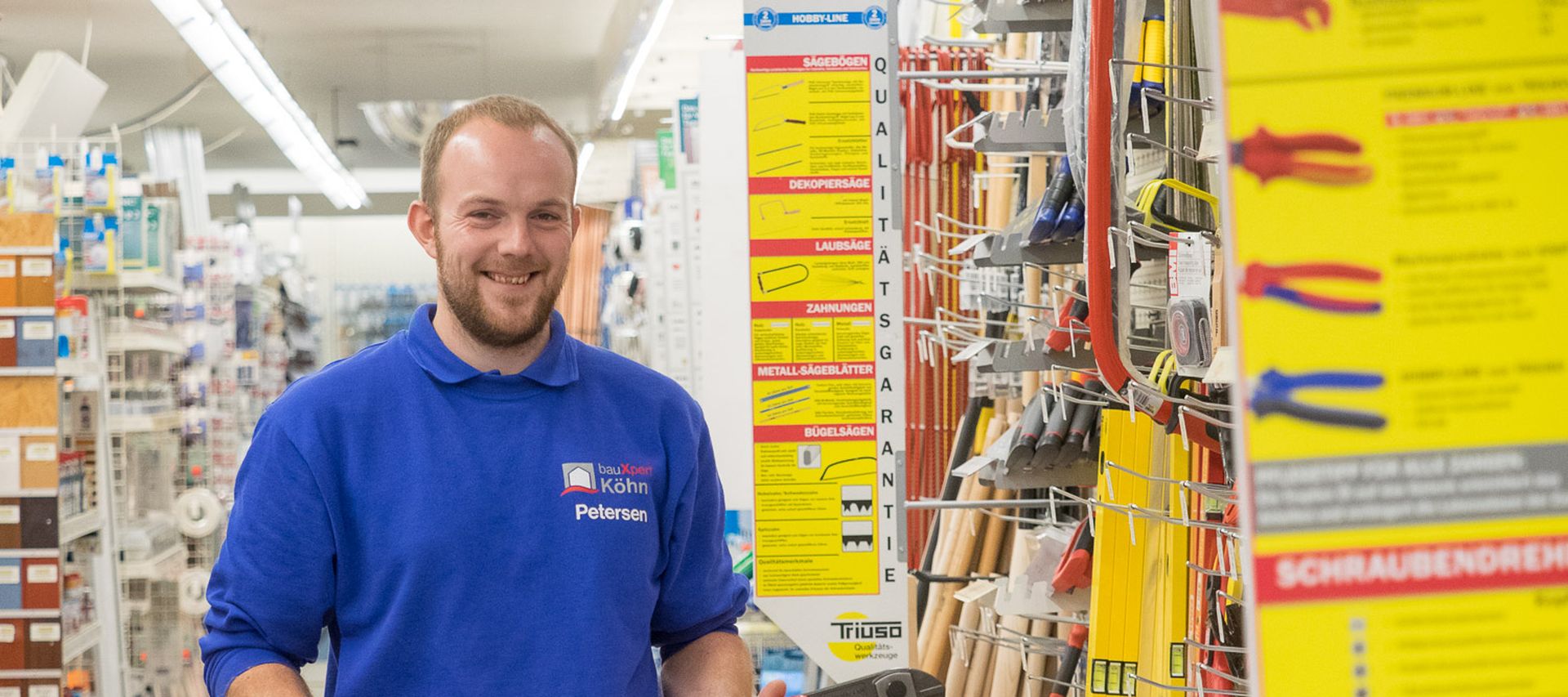 Mitarbeiter von bauXpert steht im Fachmarkt im Werkzeuggang und hält eine Bolzenschere in den Händen. Im Hintergrund sind Regale mit Handwerkzeugen und Zubehör zu sehen.