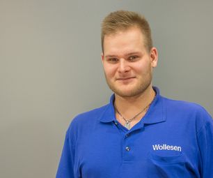 Mitarbeiter von bauXpert steht vor neutralem Hintergrund und blickt freundlich in die Kamera. Er trägt Arbeitskleidung mit Namenszug.