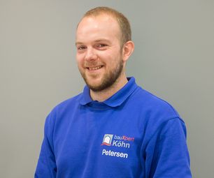 Mitarbeiter von bauXpert steht vor neutralem Hintergrund und lächelt leicht in die Kamera. Er trägt Arbeitskleidung mit Namenszug und Firmenlogo.