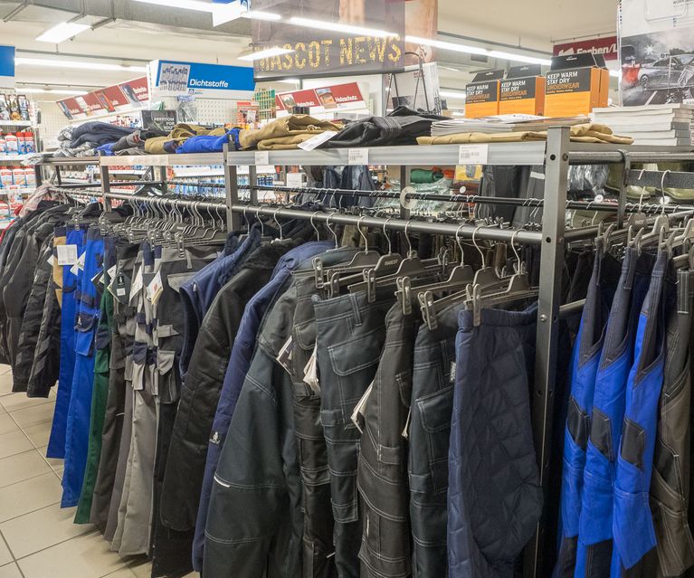 Arbeitskleidung wie Arbeitshosen und Jacken an Kleiderständern im Arbeitsschutzbereich eines bauXpert Fachmarkts.