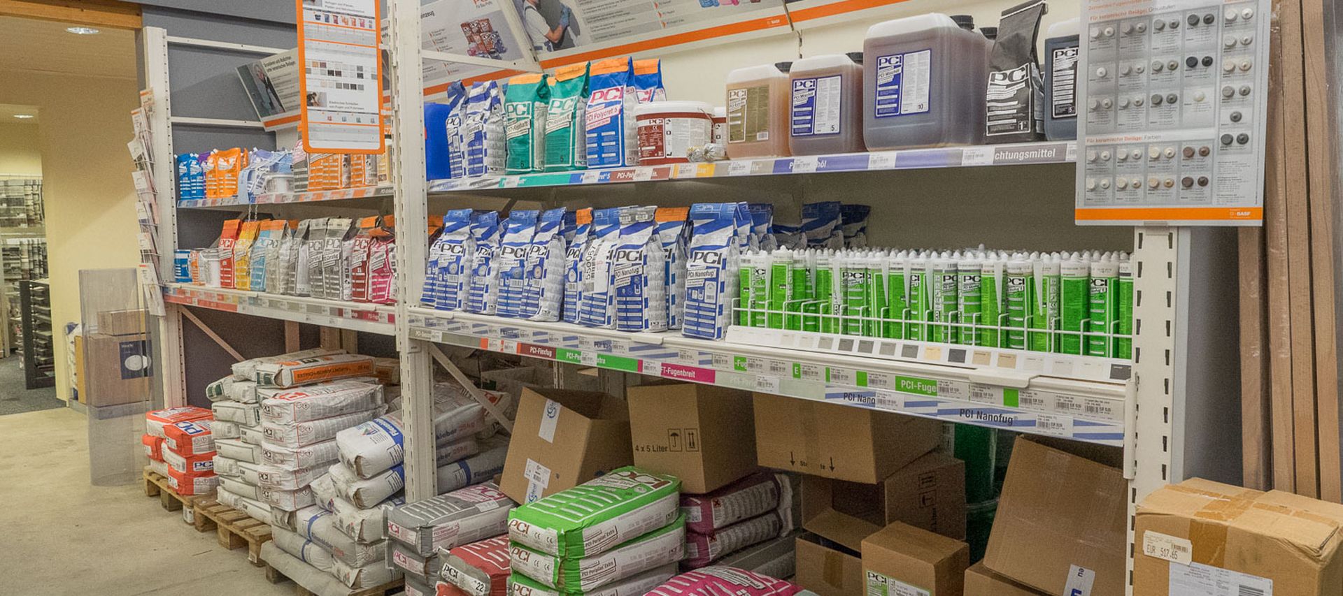 Regalfläche im Fachmarkt mit Bauchemie von PCI, darunter Kleber, Fugenmörtel, Dichtstoffe und Zubehör für Fliesen- und Abdichtungsarbeiten. Die Produkte sind nach Anwendung sortiert.