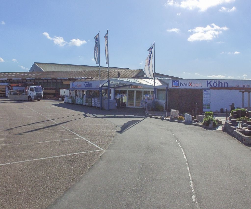 Gesamtansicht des bauXpert Standorts Köhn mit Einfahrt, Eingangsbereich und angrenzender Gartenausstellung bei sonnigem Wetter. Auf dem Gelände sind Lagerflächen, Fahrzeuge und Musterbereiche zu sehen.