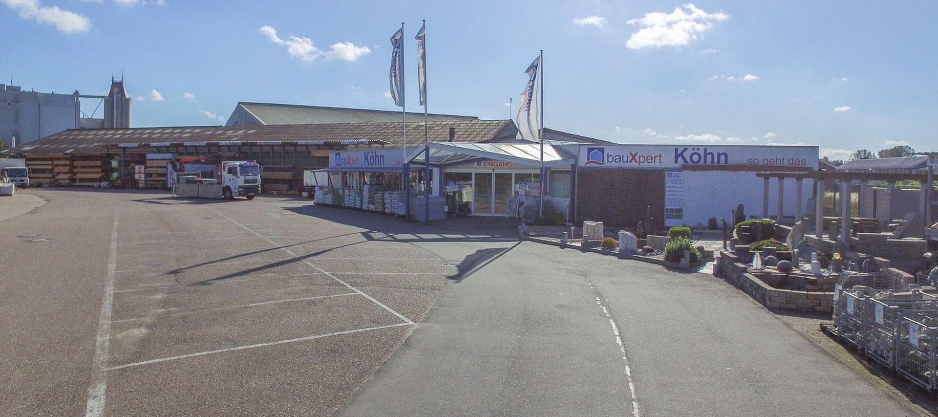 Gesamtansicht des bauXpert Standorts Köhn mit Einfahrt, Eingangsbereich und angrenzender Gartenausstellung bei sonnigem Wetter. Auf dem Gelände sind Lagerflächen, Fahrzeuge und Musterbereiche zu sehen.