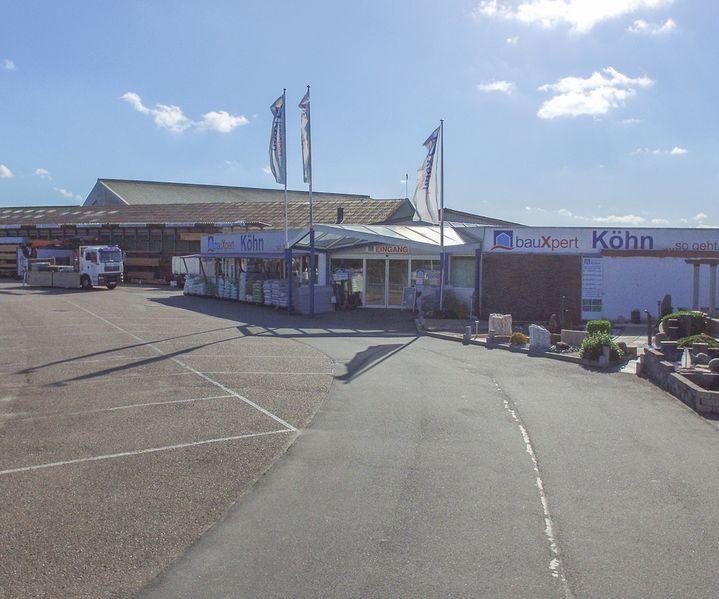 Gesamtansicht des bauXpert Standorts Köhn mit Einfahrt, Eingangsbereich und angrenzender Gartenausstellung bei sonnigem Wetter. Auf dem Gelände sind Lagerflächen, Fahrzeuge und Musterbereiche zu sehen.