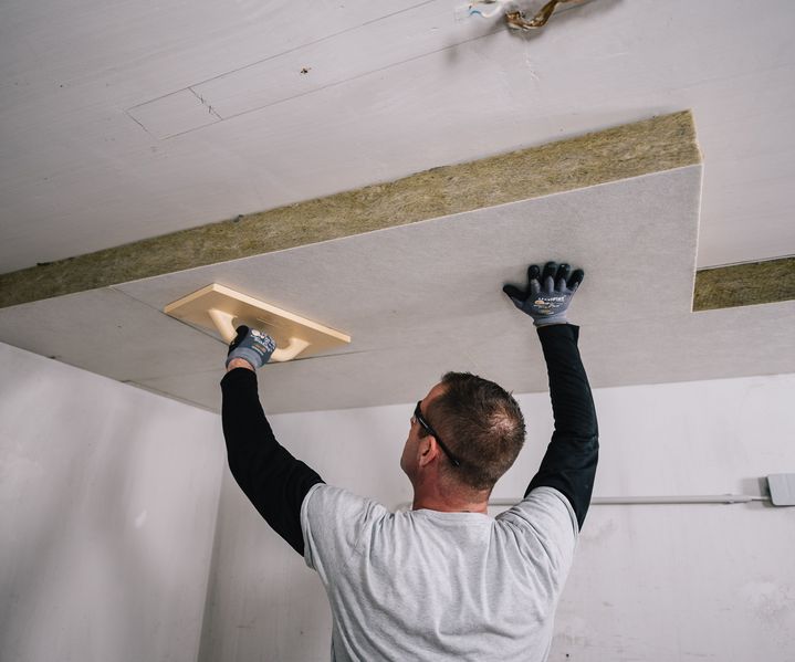 Handwerker befestigt Dämmplatten an der Kellerdecke und glättet die Oberfläche mit einem Werkzeug. Die Platten liegen flächig an.