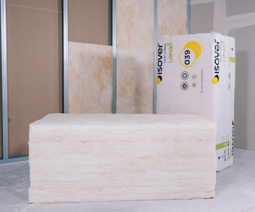 Dämmmatten liegen gestapelt vor einer Wandkonstruktion, daneben eine verpackte Rolle Dämmstoff. Die Struktur des Materials ist gut sichtbar.