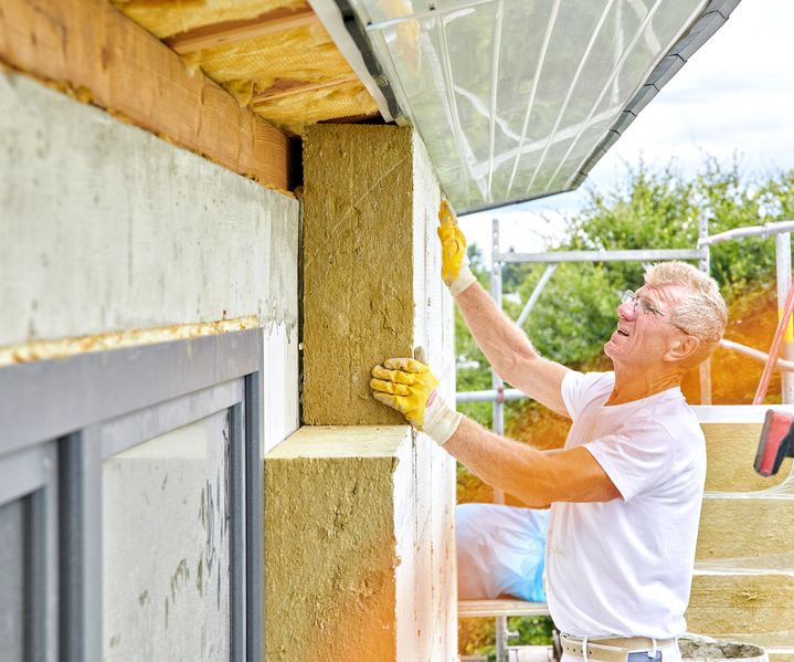 Handwerker bringt Dämmplatten an der Außenwand eines Gebäudes an und richtet sie unter dem Dachüberstand aus. Er arbeitet auf einem Gerüst und trägt Arbeitshandschuhe.