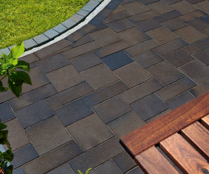 Dunkle Pflastersteine im Formatmix in Brauntönen, verlegt als Terrassen- oder Gehwegfläche mit Rasenkante und Holzbank.Pflastersteine Formatmix Titan für Terrasse und Gartenwege