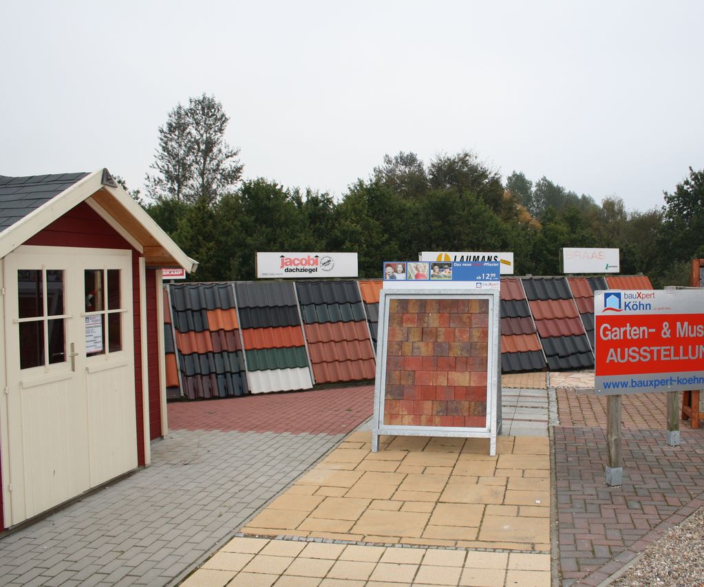 Garten- und Musterausstellung bei bauXpert mit Pflasterflächen, Dachziegelmustern und verschiedenen Materialaufbauten im Außenbereich. Hinweisschild zur Ausstellung steht im Vordergrund.