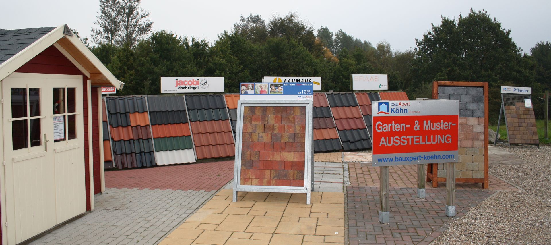 Garten- und Musterausstellung bei bauXpert mit Pflasterflächen, Dachziegelmustern und verschiedenen Materialaufbauten im Außenbereich. Hinweisschild zur Ausstellung steht im Vordergrund.