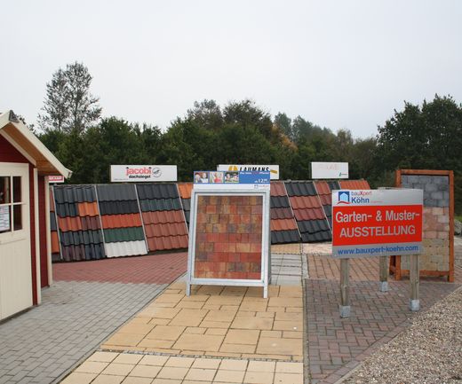 Garten- und Musterausstellung bei bauXpert mit Pflasterflächen, Dachziegelmustern und verschiedenen Materialaufbauten im Außenbereich. Hinweisschild zur Ausstellung steht im Vordergrund.