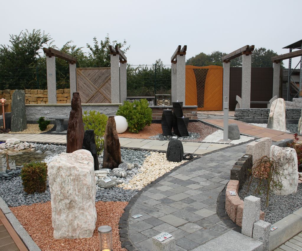 Gartenausstellung bei bauXpert mit angelegten Wegen, Zierkiesflächen, Natursteinen und Gestaltungselementen wie Findlingen und Wasserspielen. Die Musterflächen zeigen unterschiedliche Materialien und Verlegearten.