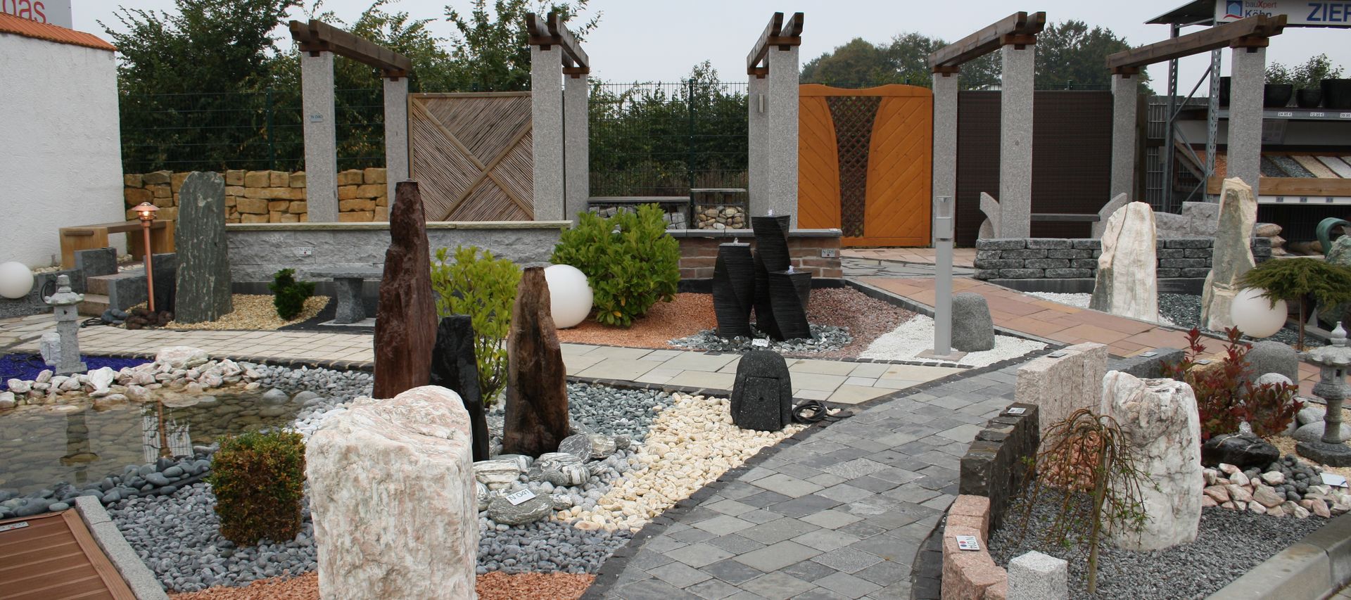 Gartenausstellung bei bauXpert mit angelegten Wegen, Zierkiesflächen, Natursteinen und Gestaltungselementen wie Findlingen und Wasserspielen. Die Musterflächen zeigen unterschiedliche Materialien und Verlegearten.