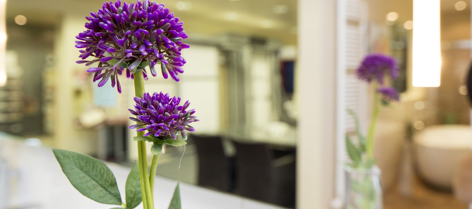 Violette Zierblume in einer Vase auf einem weißen Waschtisch, im Hintergrund ein unscharf dargestellter Badezimmer-Ausstellungsbereich.