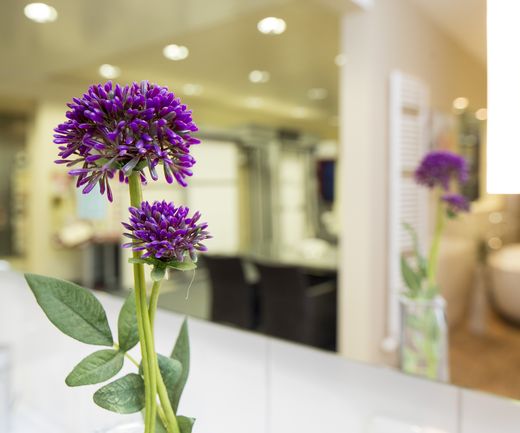Violette Zierblume in einer Vase auf einem weißen Waschtisch, im Hintergrund ein unscharf dargestellter Badezimmer-Ausstellungsbereich.